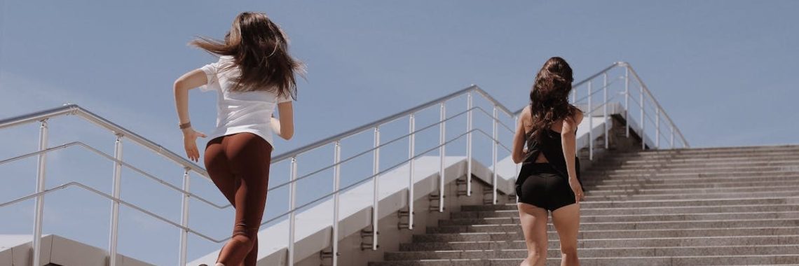 Two women running up outdoor stairs for fitness under a clear blue sky.